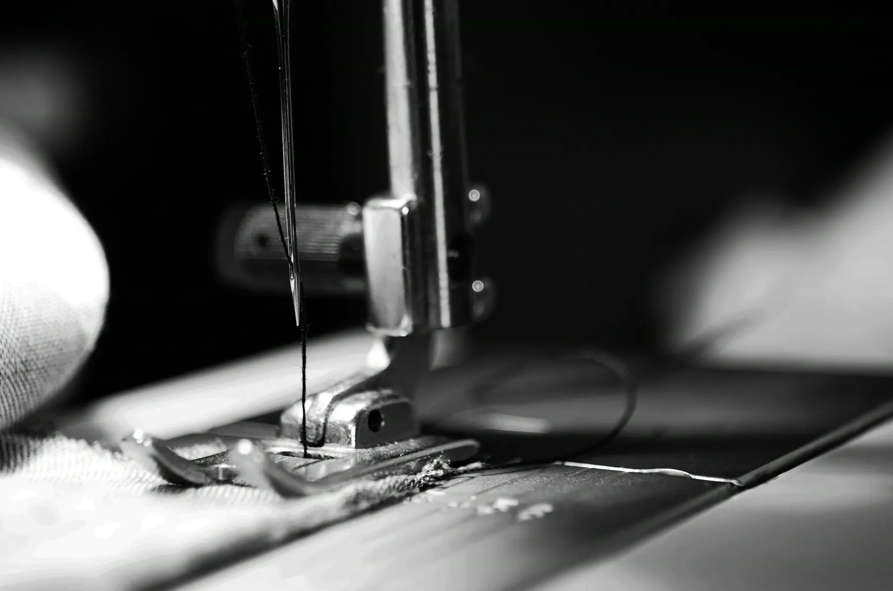 Black and white close-up of a sewing machine needle and presser foot stitching cloth, thread trailing
